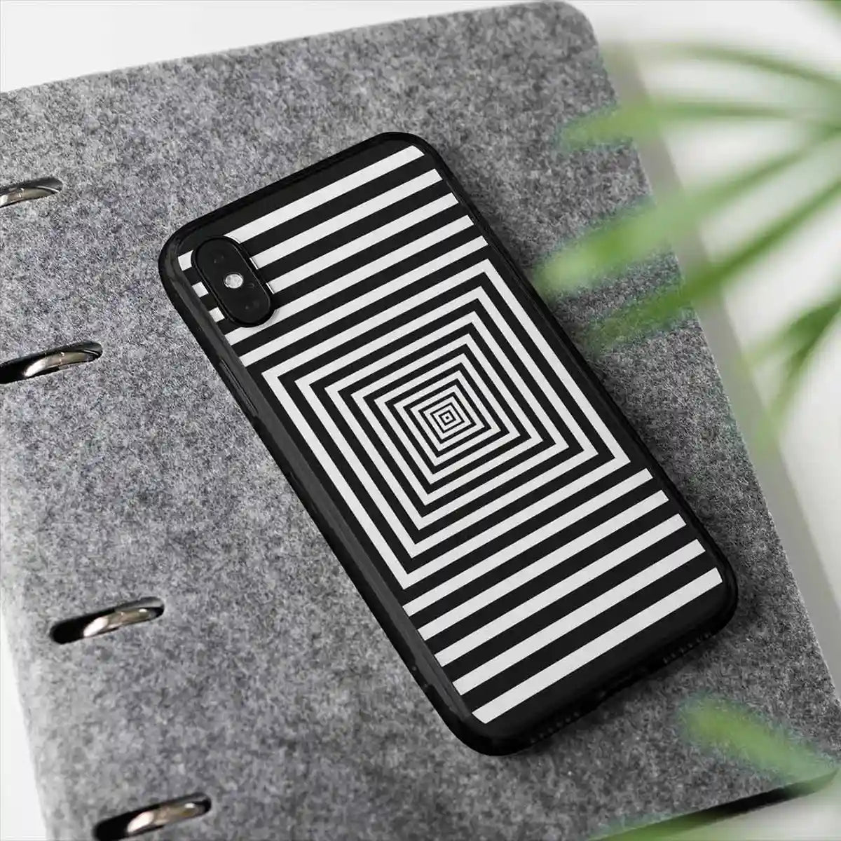 Squares digital art print on a clear phone case placed by a plant, featuring bold concentric black-and-white squares, adding a modern and artistic touch to the scene.